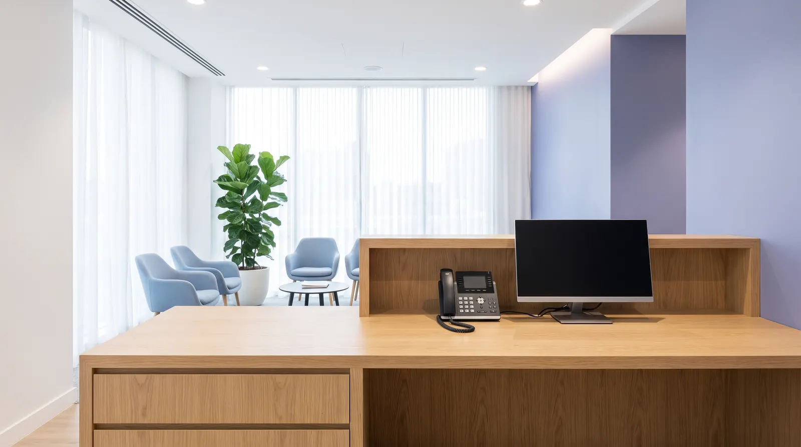 Mostrador de recepción de una clínica con el teléfono y la pantalla de agenda, y la sala de espera al fondo