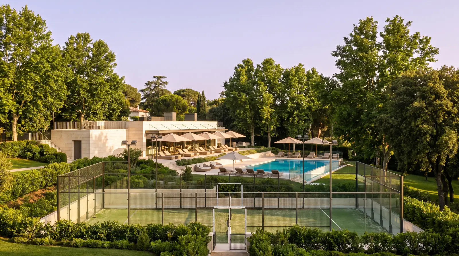 Instalaciones de un club deportivo con pistas de pádel acristaladas, la terraza de la casa club y la piscina al fondo