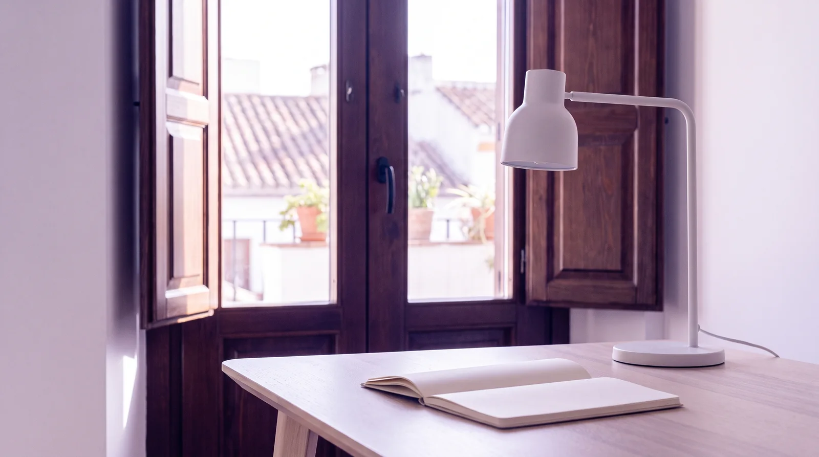 Oficina luminosa con una ventana de contraventanas de madera abierta a los tejados de Sevilla, con una mesa y un flexo en primer plano.
